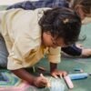 Two children drawing colorful designs on pavement with chalk on a sunny day.