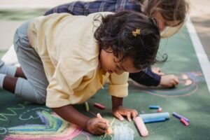 Two children drawing colorful designs on pavement with chalk on a sunny day.