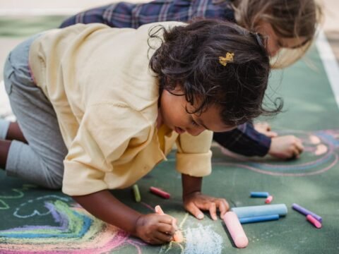 Two children drawing colorful designs on pavement with chalk on a sunny day.