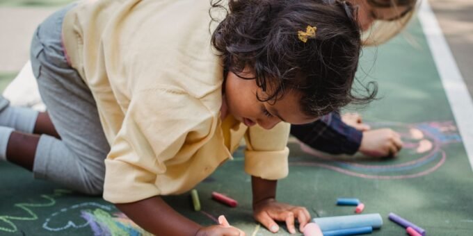 Two children drawing colorful designs on pavement with chalk on a sunny day.